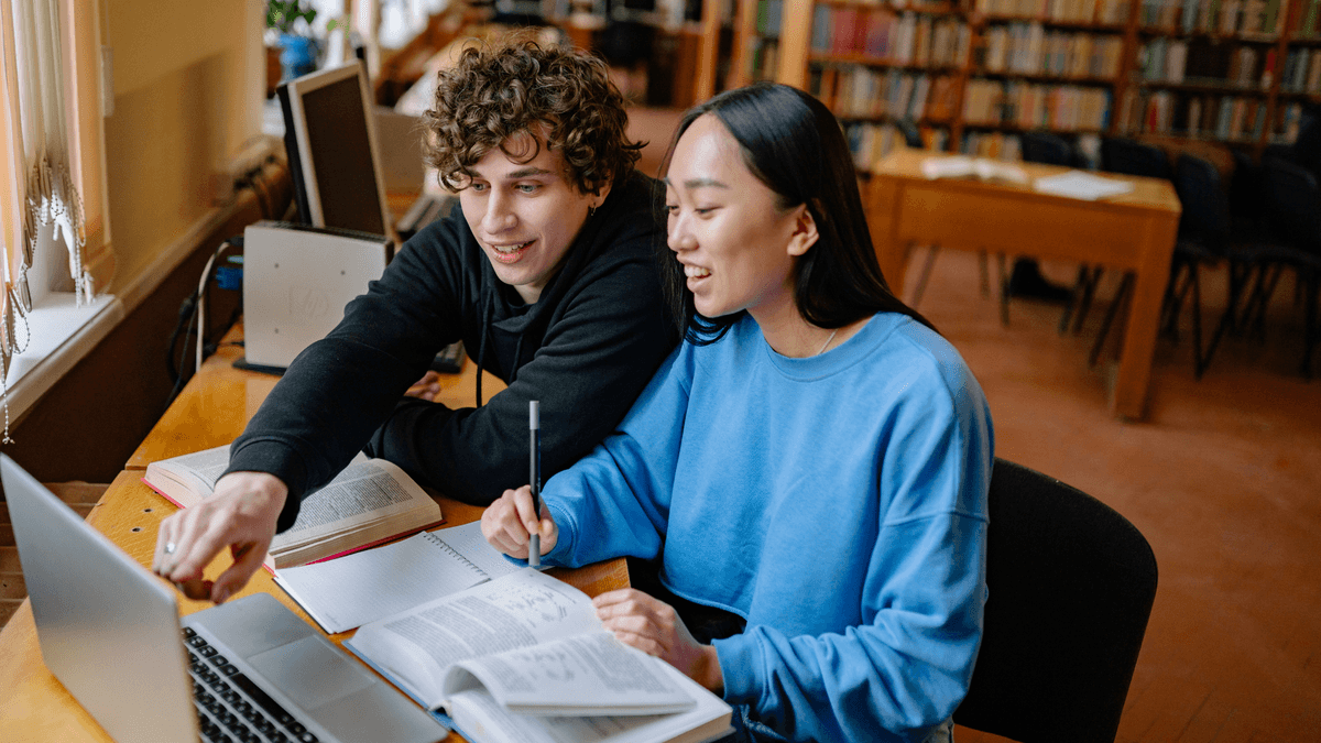 Two students sitting in a library, looking at a laptop screen and open textbooks together.