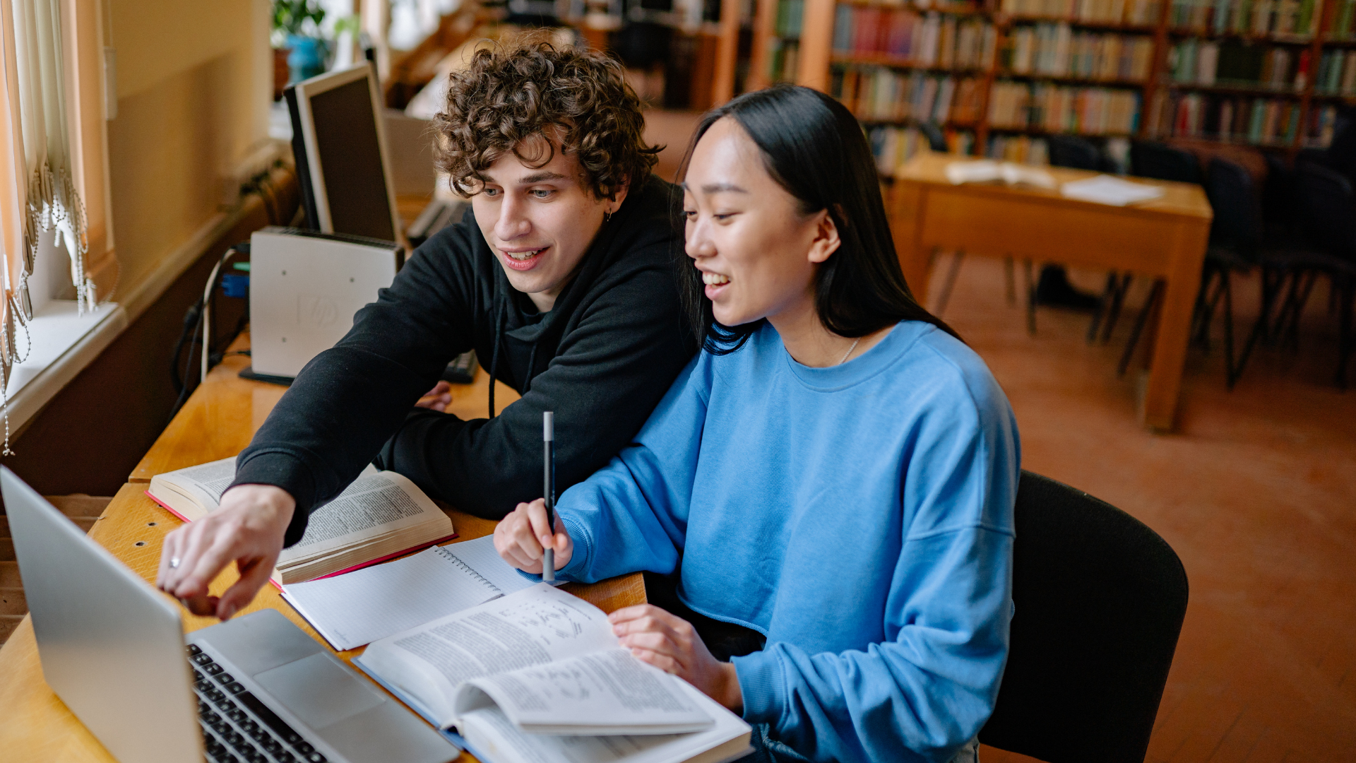 Two students sitting in a library, looking at a laptop screen and open textbooks together.