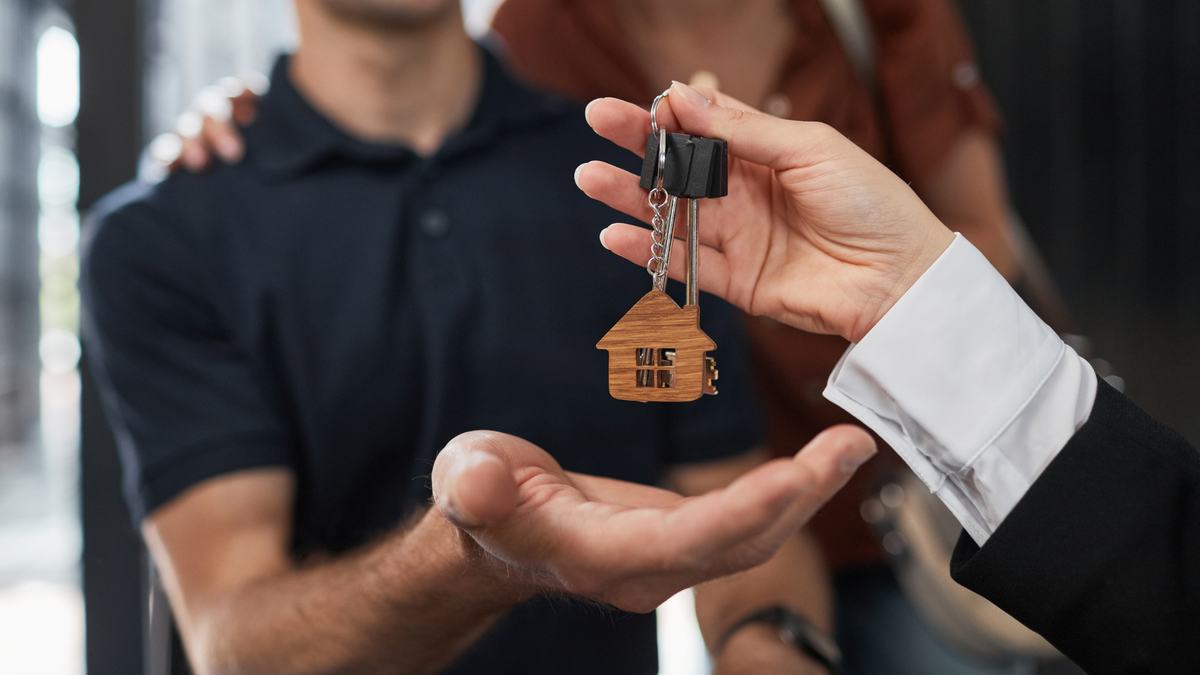 Real estate agent handing keys with a wooden house-shaped keychain to someone signifying a first home buyer