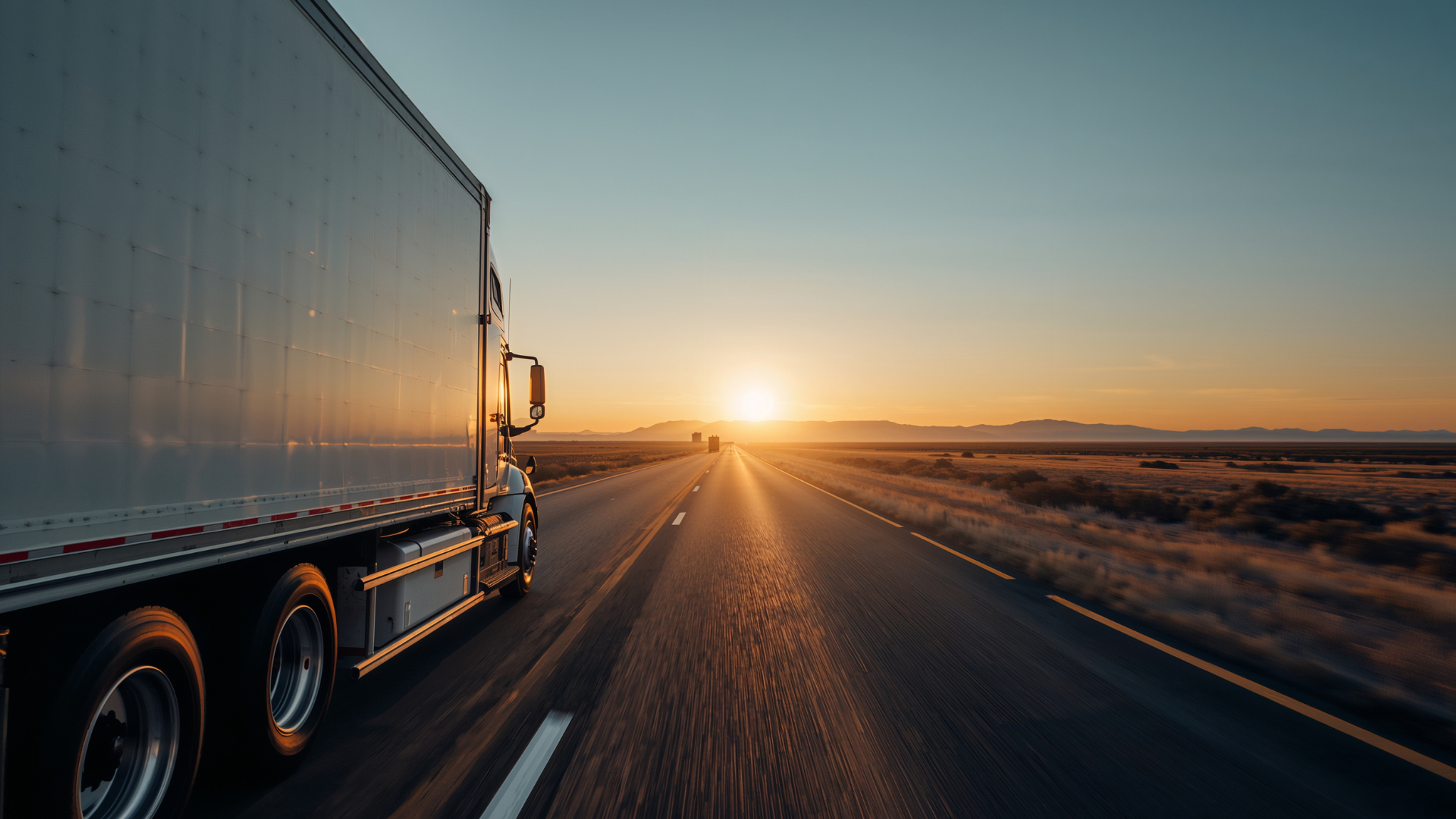 White semi-truck drives down a long highway toward a bright sunset on the horizon